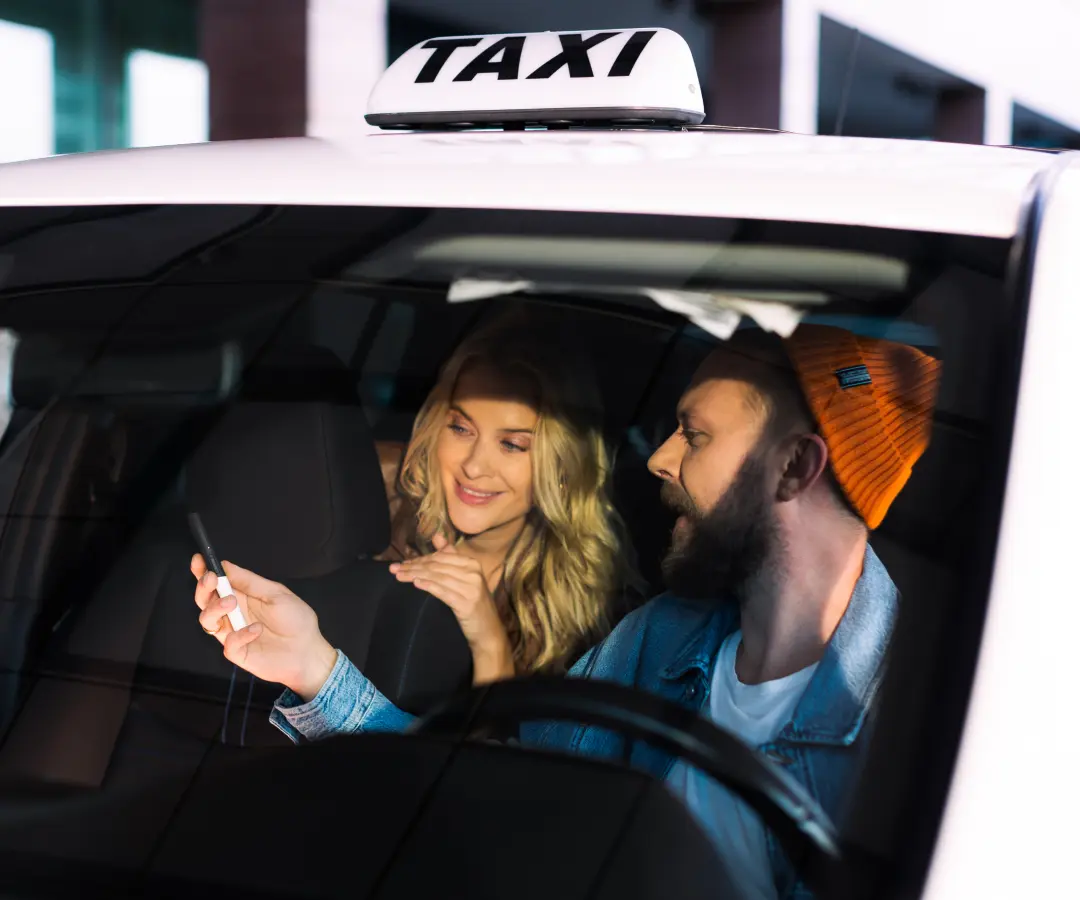 Muž sedí na sedadle vodiča taxíka a ukazuje niečo žene na zadnom sedadle na prekladači, ktorý drží v ruke. Žena sa so záujmom usmieva. Na streche auta je vidieť nápis taxi.