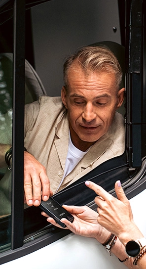 Muž sedí v aute a hovorí do zariadenia Vasco Q1, ktoré mu podáva iná osoba.