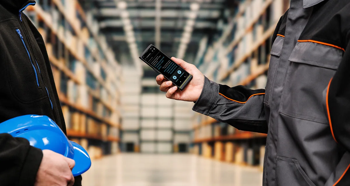 Dvaja ľudia stoja oproti sebe; jeden drží prilbu a druhý Vasco Translator V4. Na obrazovke zariadenia je aktívne rozhranie hlasového prekladu.