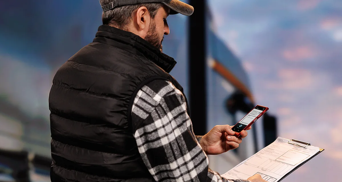 Vodič kamióna stojí pred vozidlom, v jednej ruke drží dokumenty a v druhej Vasco Translator V4. Na displeji zariadenia je zobrazená funkcia prekladu z fotografie.