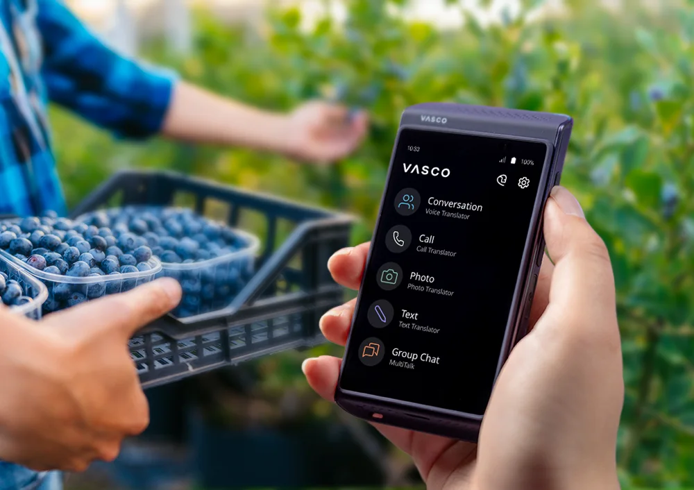 Vasco Translator Q1 v popredí, v pozadí kríky čučoriedok a osoba držiaca prepravku so zberom.