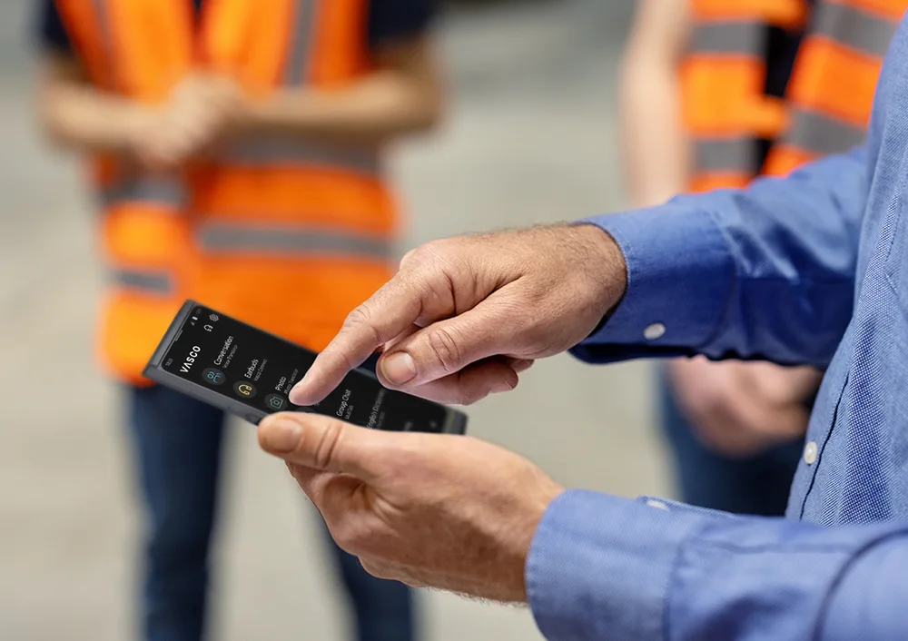 Osoba drží Vasco V4, pričom v pozadí sú viditeľní ľudia v pracovnom oblečení.