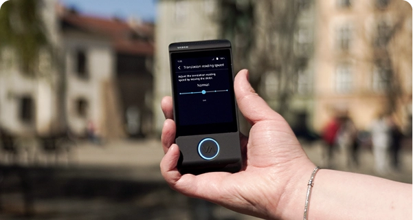 Záber zblízka na ruku osoby, ktorá drží čierny elektronický prekladač Vasco, na rozmazanom exteriérovom pozadí mestského námestia s historickými budovami. Na obrazovke zariadenia sú zobrazené nastavenia „Rýchlosť čítania prekladu“ s posuvníkom nastaveným na „Normálne“ a poznámkou: „Rýchlosť čítania prekladu nastavte posunutím posuvníka.“ Osoba držiaca zariadenie má na ruke strieborný náramok. Jasné denné svetlo osvetľuje rozhranie obrazovky, vďaka čomu sú nastavenia dobre viditeľné. 